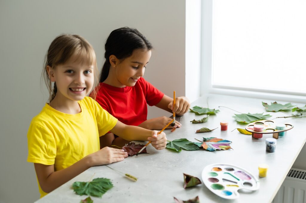 two girls paint autumn leaves at table.